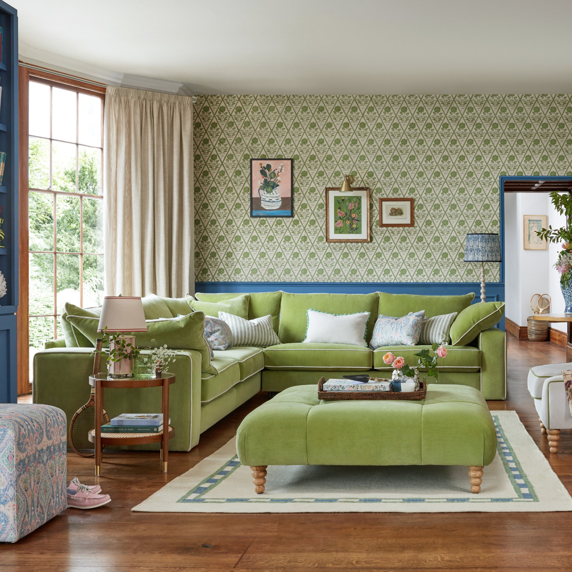 A living room with a corner green velvet sofa with contrasting white piping set against a green print wallpaper on the wall