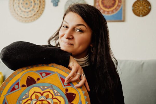 A woman poses with a colorful mandala art piece in a cozy indoor setting.