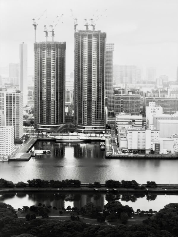 A black-and-white cityscape showing tall skyscrapers under construction with cranes on top, surrounded by other buildings, a river reflecting the skyline, and a tree-lined park in the foreground.