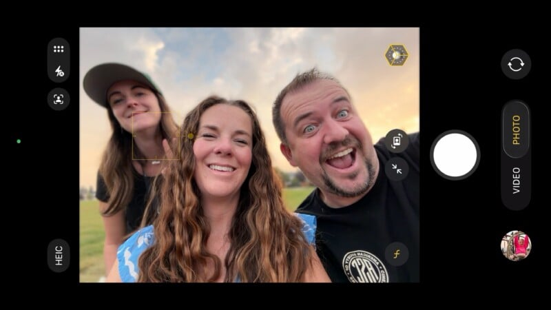 A smartphone camera app interface shows three smiling adults outdoors posing for a selfie at sunset, with the sky and grass in the background. The camera settings and shutter button are visible on screen.