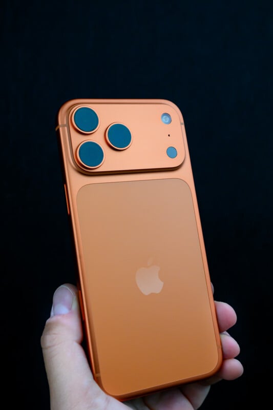 A hand holds an orange smartphone with three rear cameras and the Apple logo visible against a dark background.