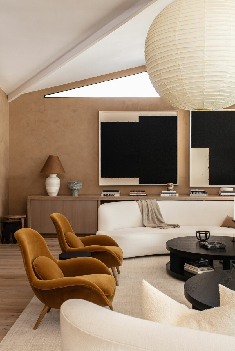 Modern living room with mustard velvet chairs, white curved sofas, round black coffee table, paper lantern pendant, and neutral tan walls with abstract black and white art above a console.