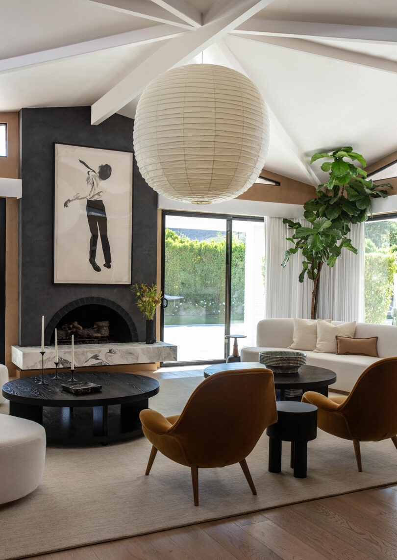 Modern living room with mustard yellow armchairs, a large paper lantern light, a black round coffee table, artwork above a fireplace, and a tall indoor plant by floor-to-ceiling windows.
