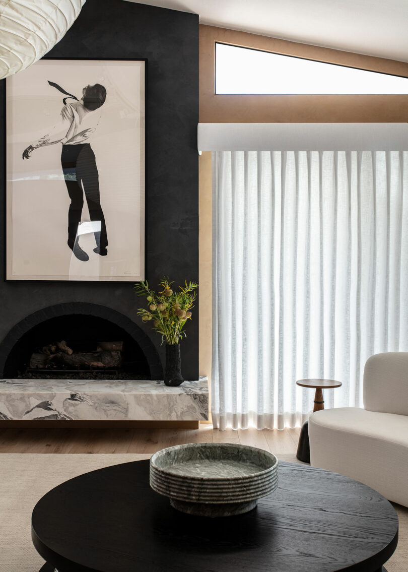 Modern living room with a black fireplace, abstract art above the mantel, light curtains, a white armchair, and a round dark coffee table with a ceramic bowl.