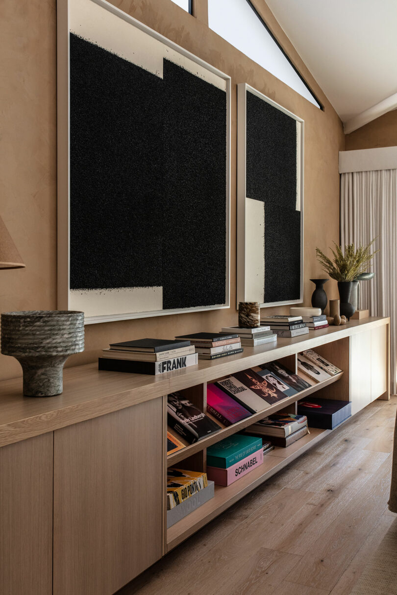 A modern living room with a long wooden sideboard, large abstract black artworks above, books and decorative items arranged neatly on top and shelves below.