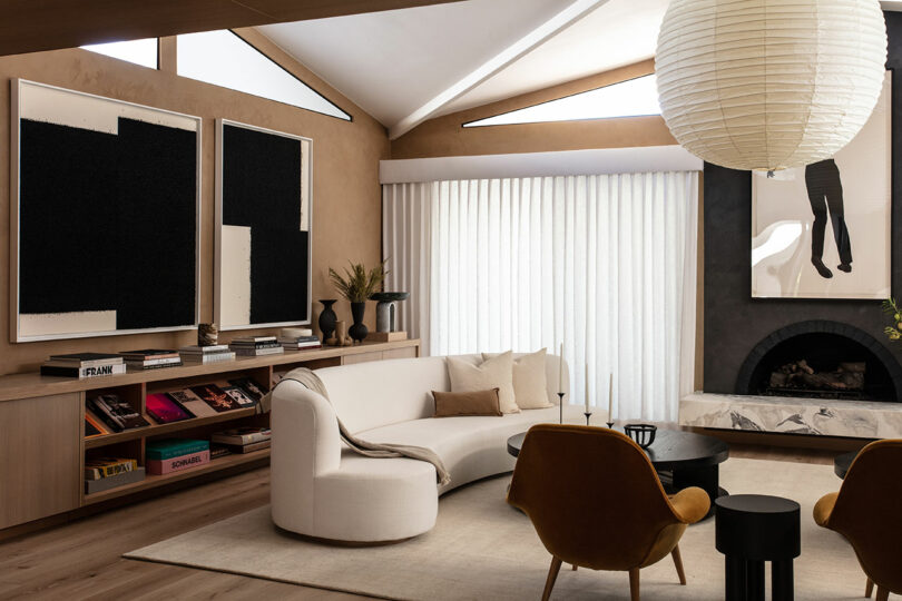 Modern living room with a curved white sofa, two mustard armchairs, black round coffee table, paper lantern light, neutral decor, bookshelves, and abstract art on the walls.