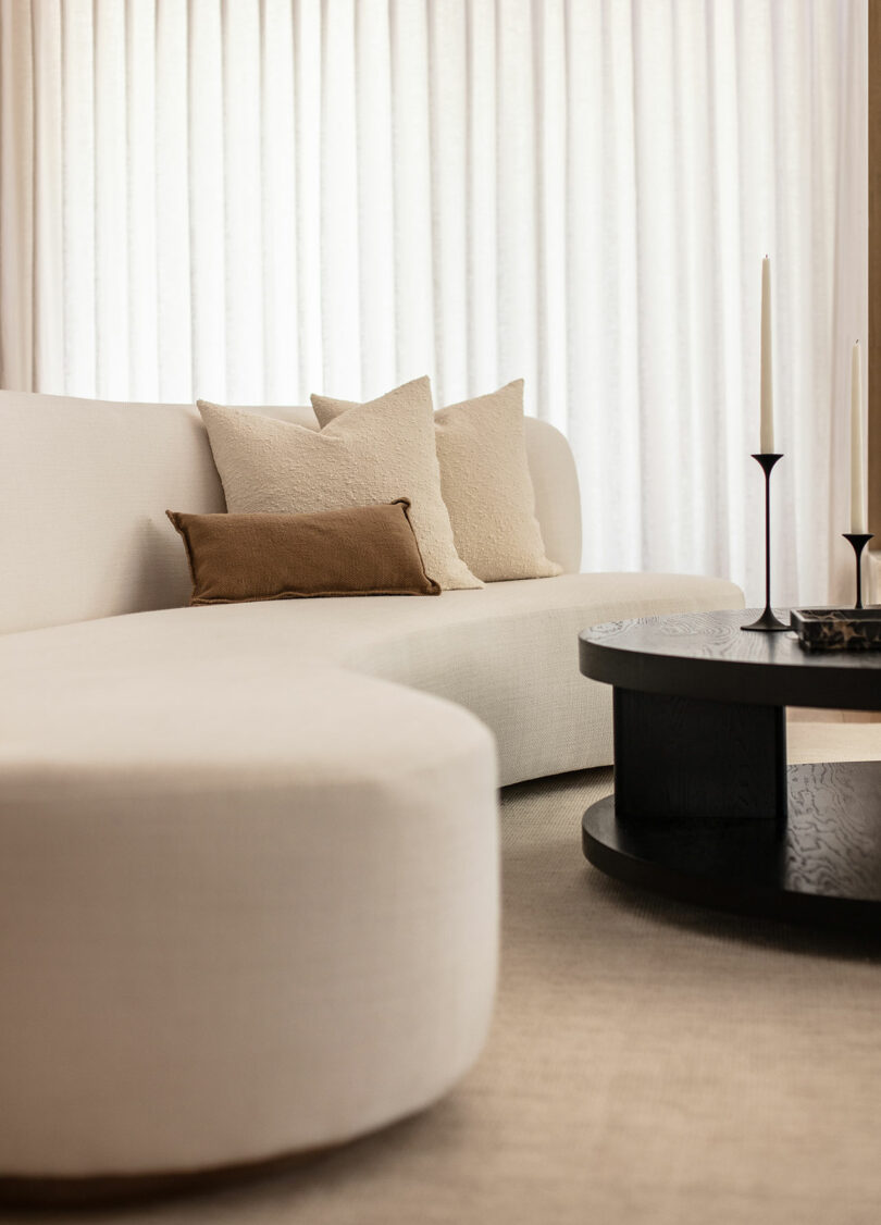 Curved white sofa with neutral cushions next to a round black coffee table with two tall white candles, set against sheer white curtains.