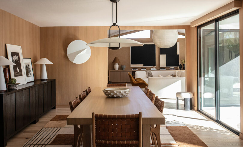 A modern dining room with a wooden table, woven chairs, sideboard, decorative lamps, and large sliding glass doors letting in natural light.