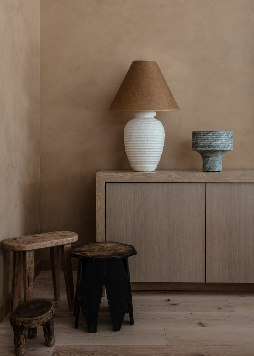 A minimalist room corner with a light wood cabinet, a white lamp with a brown shade, a green bowl, and two rustic wooden stools on a wooden floor.