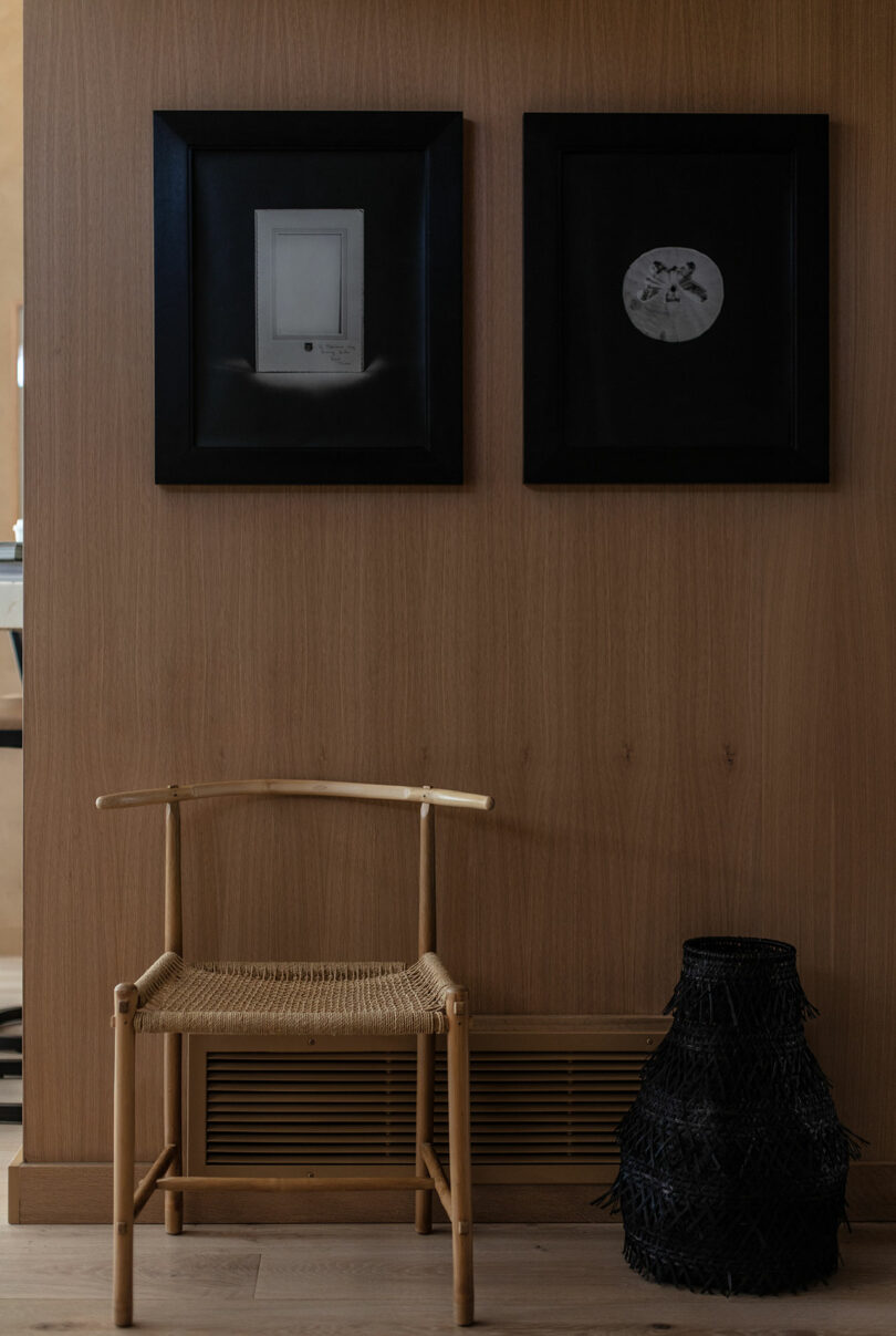 A wooden chair sits in front of a vent below two framed artworks on a wood-paneled wall, with a black woven basket on the floor to the right.