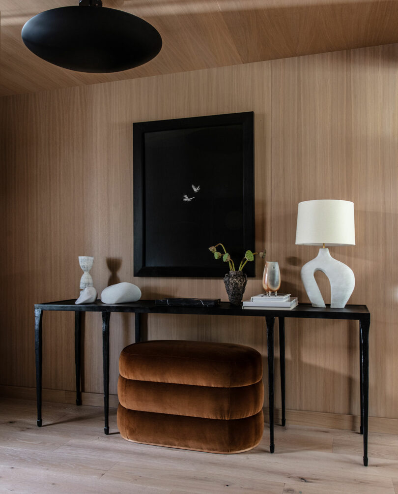 A modern console table with sculptural decor, a lamp, and a vase sits against a wood-paneled wall, with a brown velvet ottoman and a black-framed artwork above.