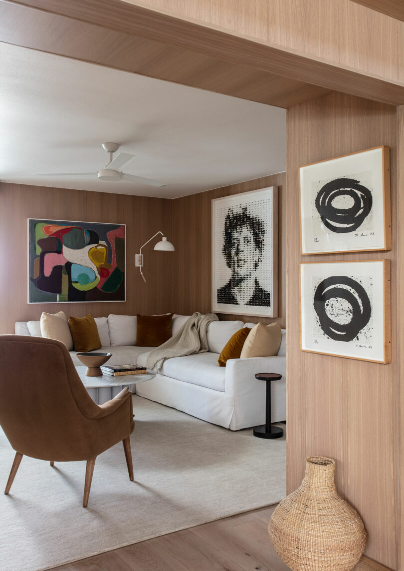 A modern living room with wood-paneled walls, white sectional sofa, brown chair, abstract and portrait artwork, small tables, and a woven basket on the floor.