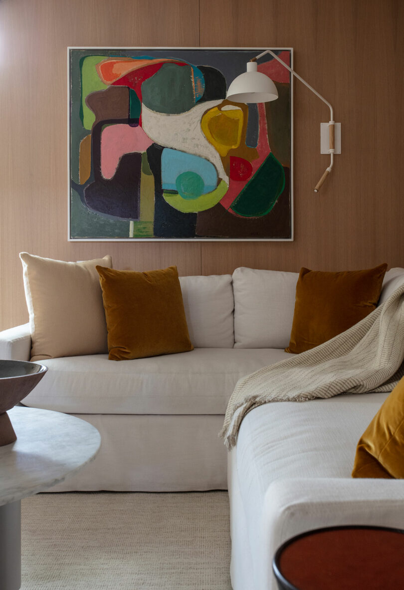 A modern living room with a white sectional sofa, mustard yellow and beige pillows, a beige throw, abstract wall art, and a white wall lamp.