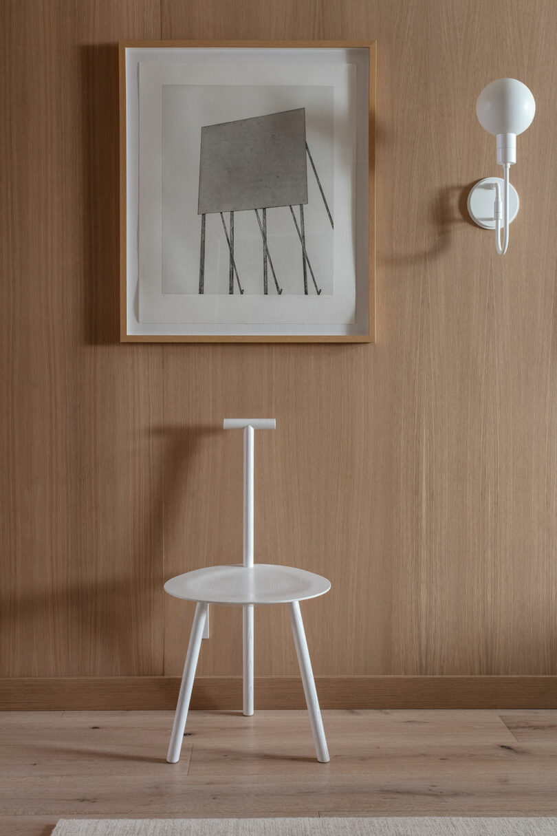 A minimalist wooden room features a framed abstract artwork, a modern white wall sconce, and a white chair with a unique backrest design.