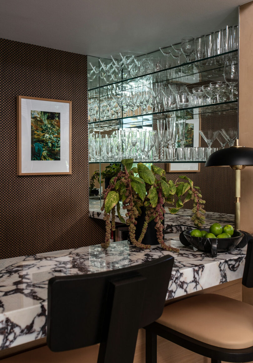 A marble dining table with black chairs sits in front of mirrored shelves filled with glassware, a leafy plant, a bowl of green apples, and a black lamp. A framed picture hangs on the wall.