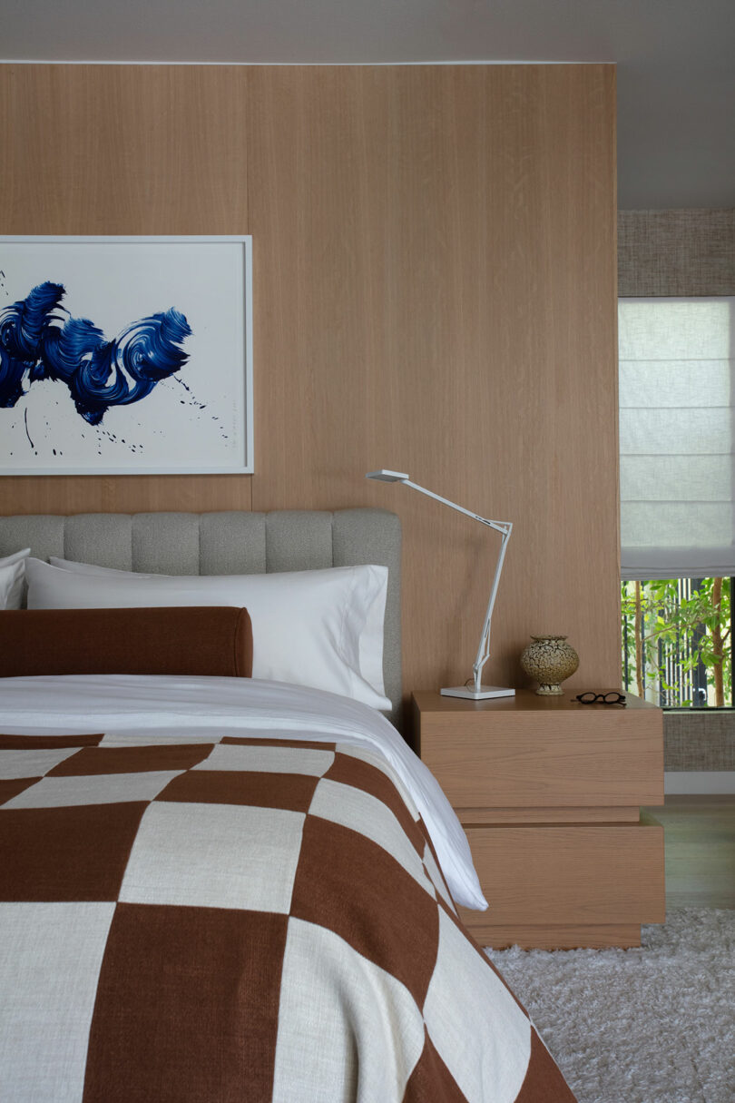 A modern bedroom with a light wood accent wall, a grey upholstered headboard, a bed with a brown and white checkered blanket, and a nightstand with a lamp and decorative items.