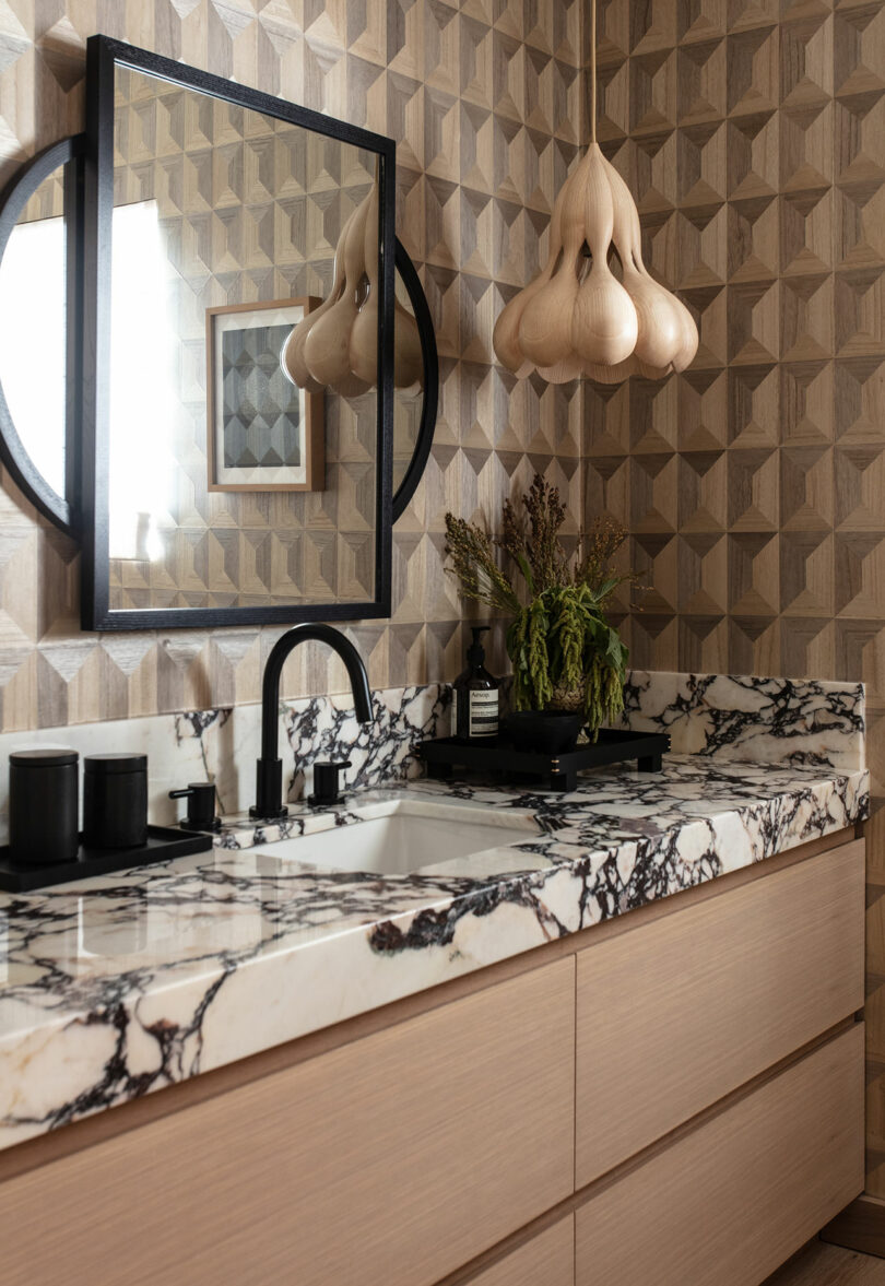 A modern bathroom with a black and white marble countertop, light wood cabinetry, geometric wall tiles, a black-framed mirror, a sculptural pendant light, and decorative plants.