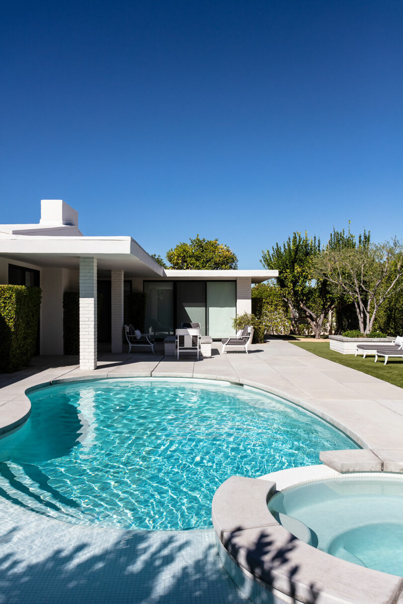 Modern house with white exterior and large windows, featuring a round swimming pool and attached spa under a clear blue sky, with patio furniture and surrounding greenery.