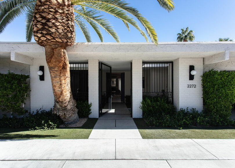 A modern, white brick building with black metal gates, a large palm tree, and the number 2272 by the entrance, surrounded by trimmed greenery.